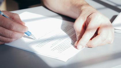 A close-up view of hands holding a blue pen and writing on a white document under bright, natural light.