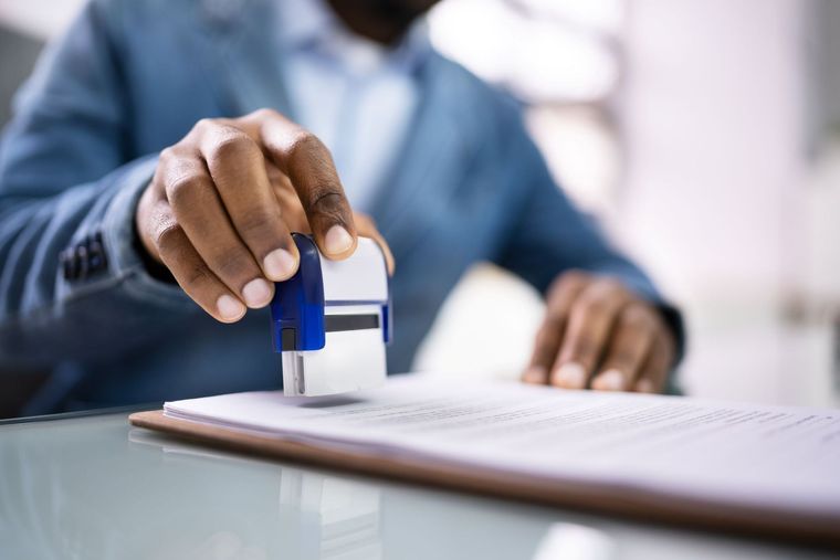 A person wearing a blue blazer uses a blue self-inking stamp to press an official seal onto a document on a desk.