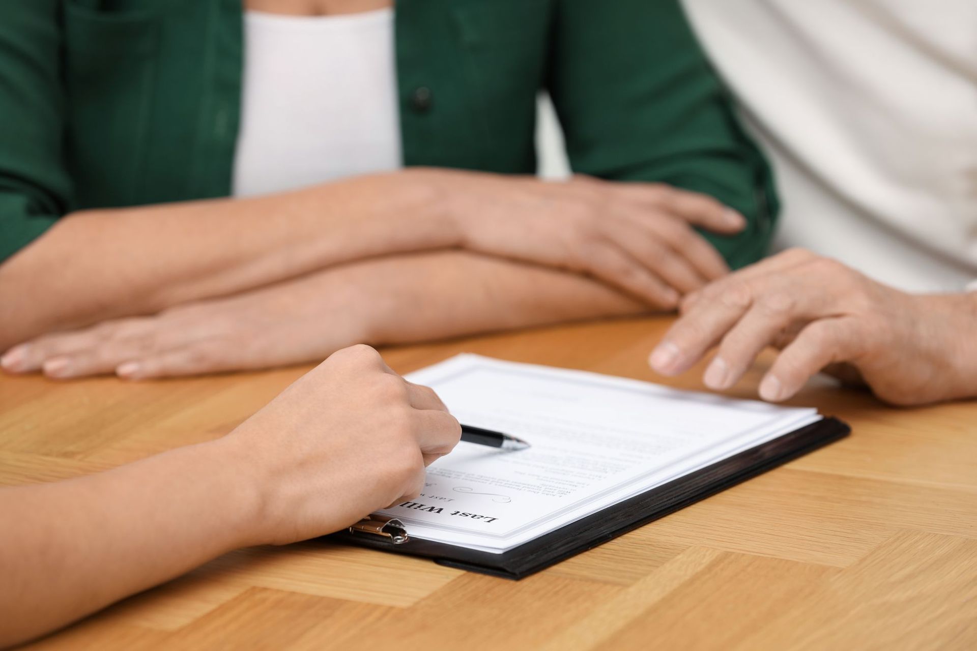 A hand holds a pen over a document on a clipboard while people sit at a wooden table.
