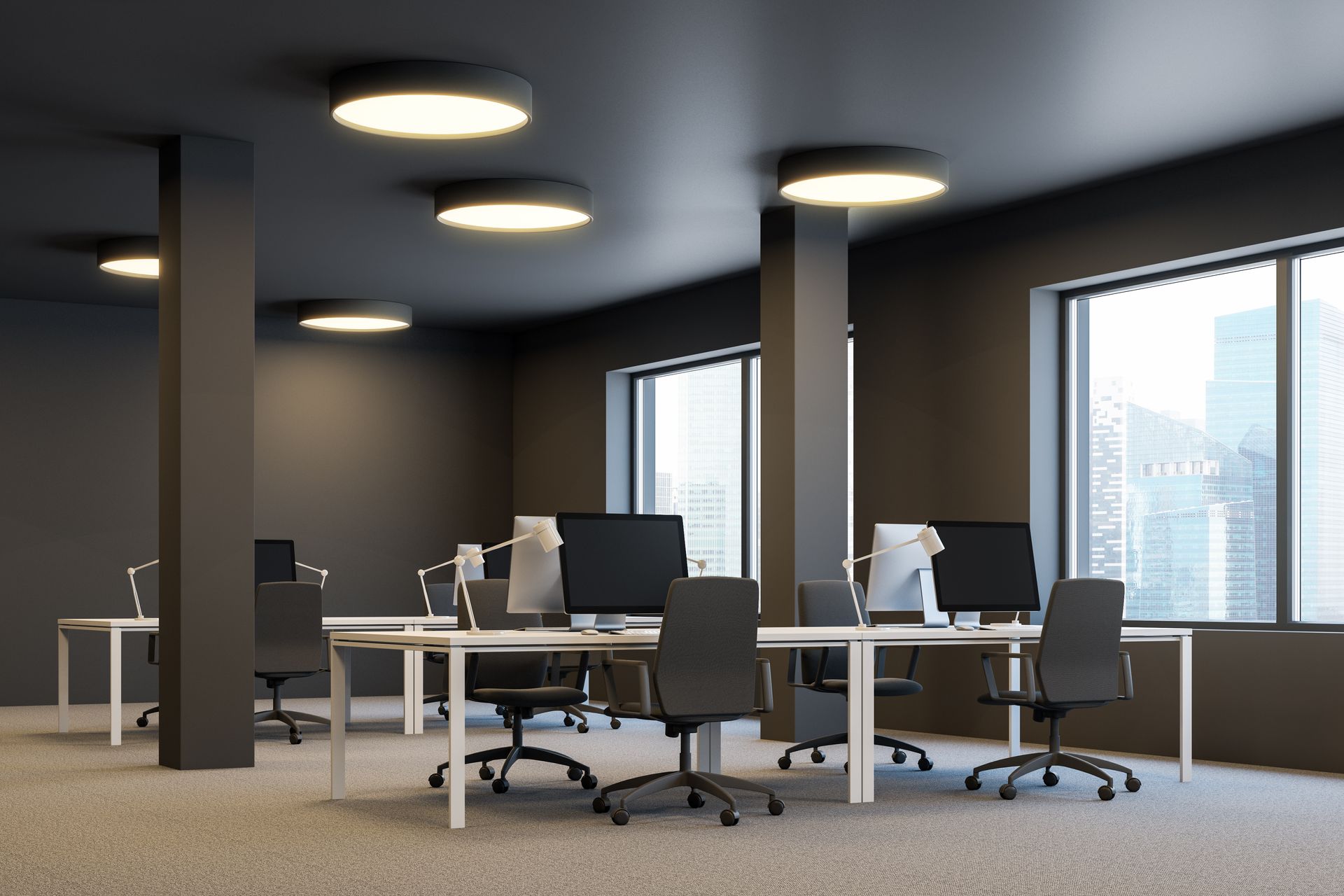 Modern open office with desks, chairs, computers, and large windows, lit by round ceiling lights.