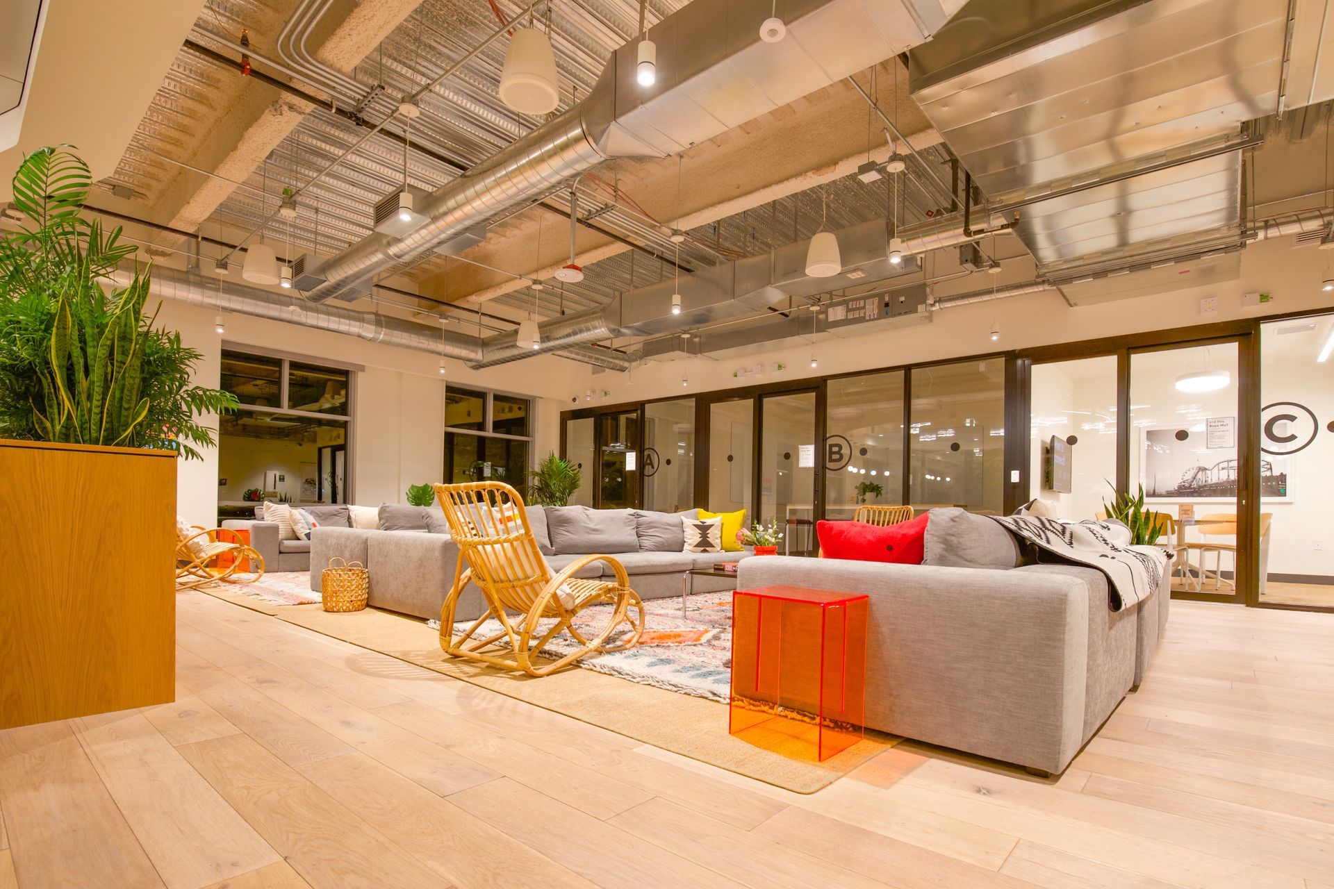 Modern office lounge with gray sofas, orange chair, wood floors, and glass walls under industrial lighting