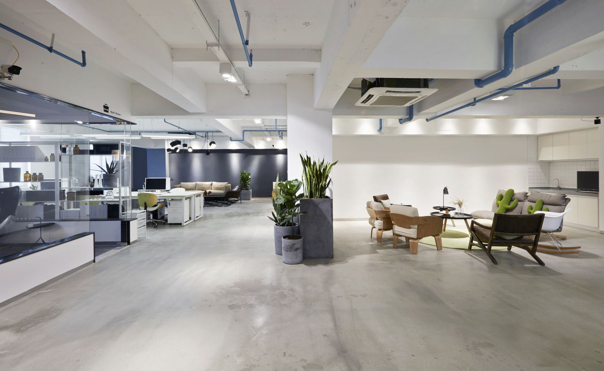 Modern open-plan office with desks, lounge seating, and concrete floors under bright ceiling lights