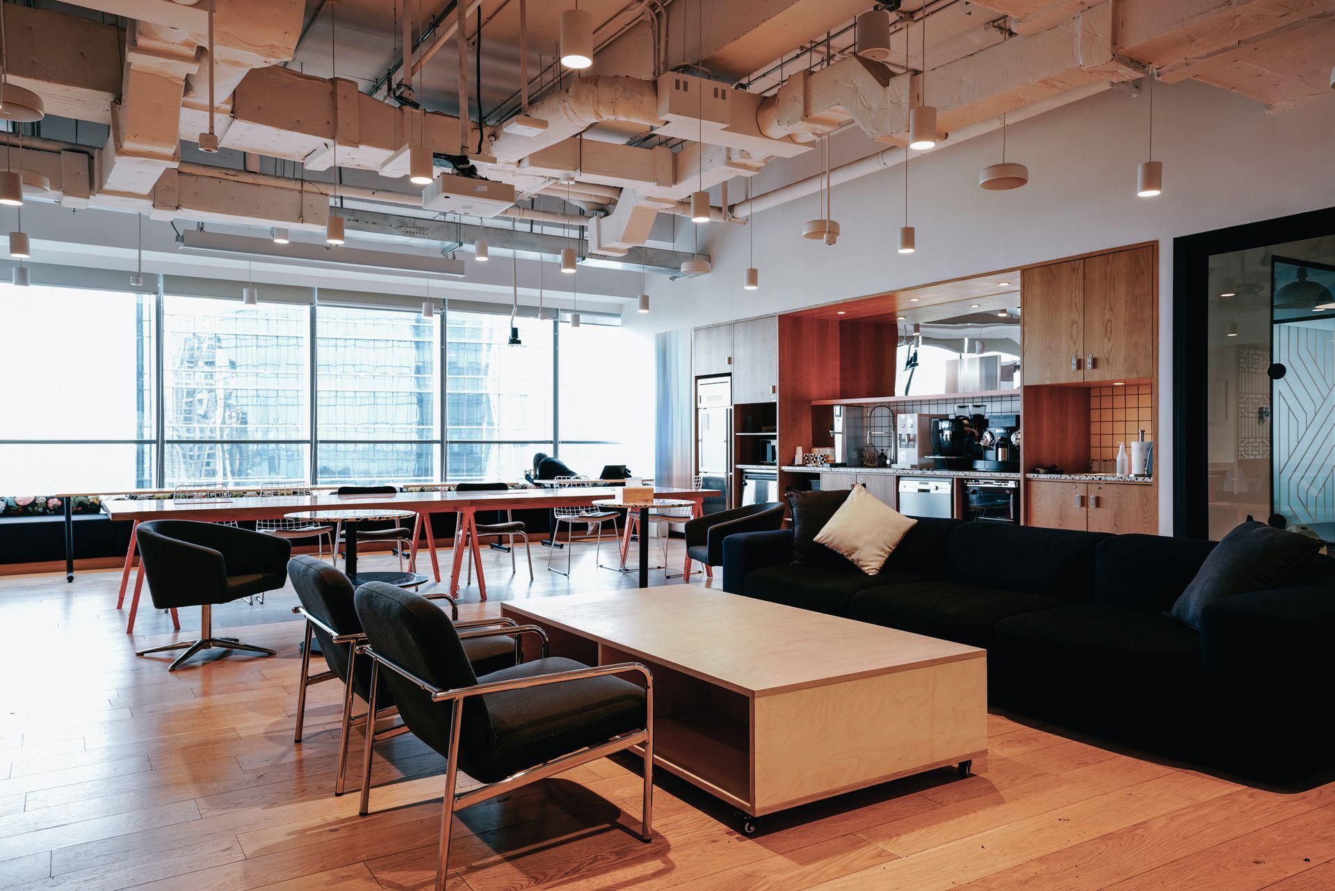 Modern office lounge with sofa, chairs, tables, and large windows under exposed ceiling beams