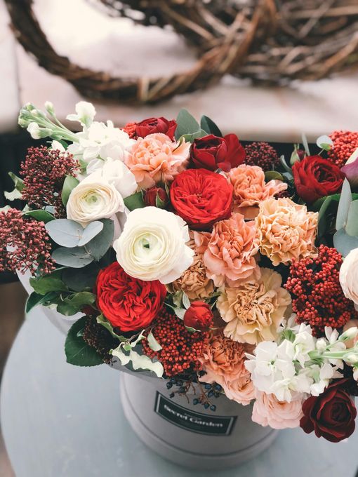A flower arrangement in a round box featuring red, peach, and white blooms with green foliage on a light surface.