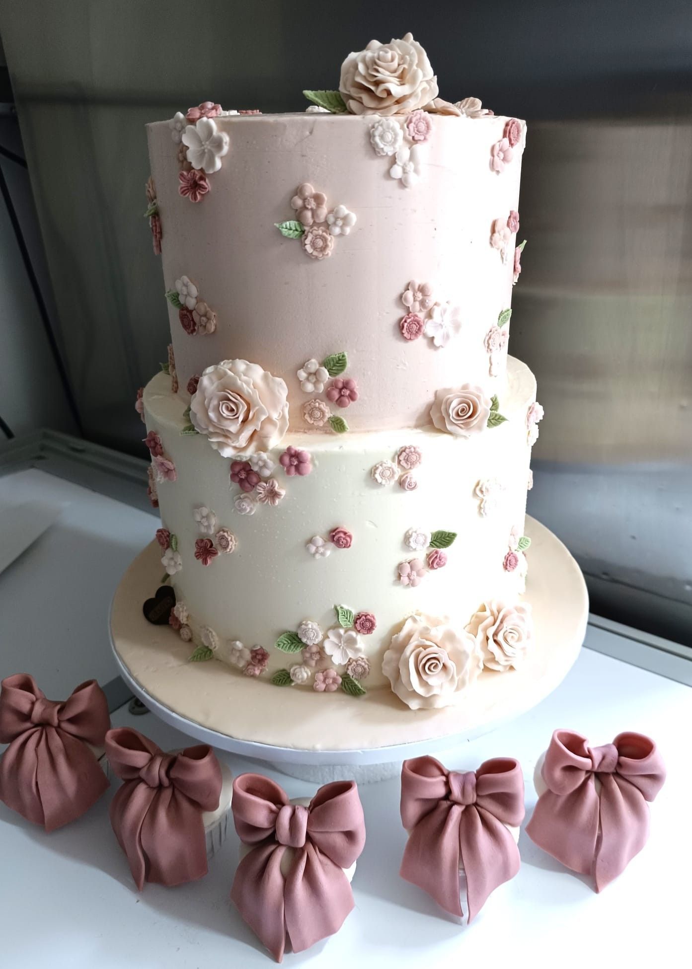 Torta a due piani rosa e bianca decorata con accenti floreali e fiocchi, presentata su un piatto bianco.