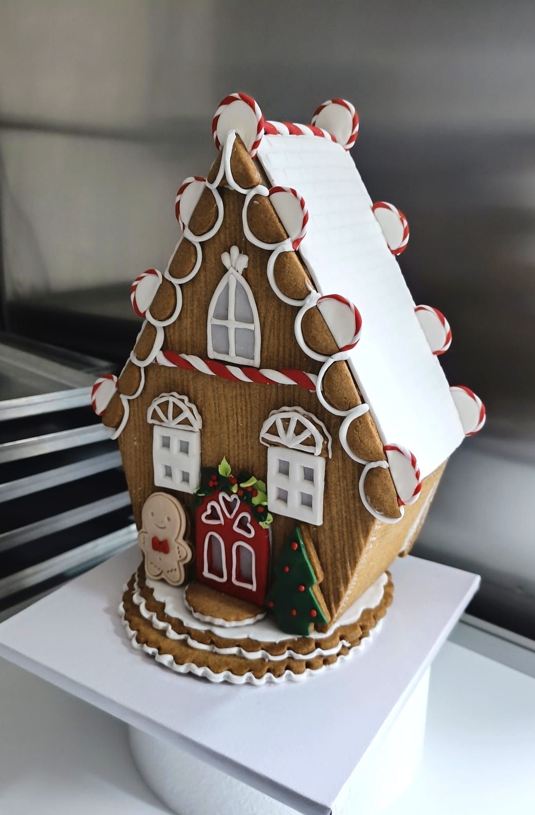 Casetta di pan di zenzero con tetto di glassa bianca, decorata con biscotti, su un'alzata per torte bianca.