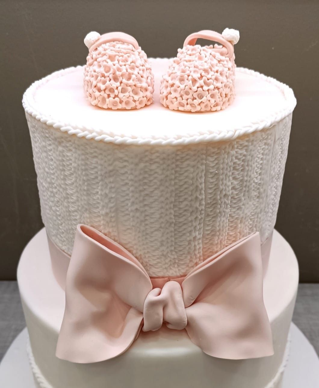 Torta per baby shower rosa e bianca con fiocco in pasta di zucchero e decorazioni a forma di scarpine per neonati lavorate a maglia.