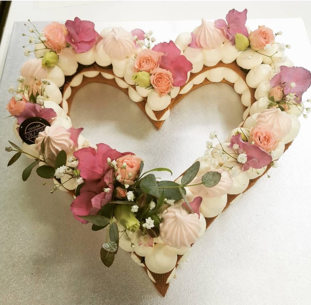 Torta a forma di cuore decorata con glassa bianca, fiori rosa e meringhe su una superficie argentata.