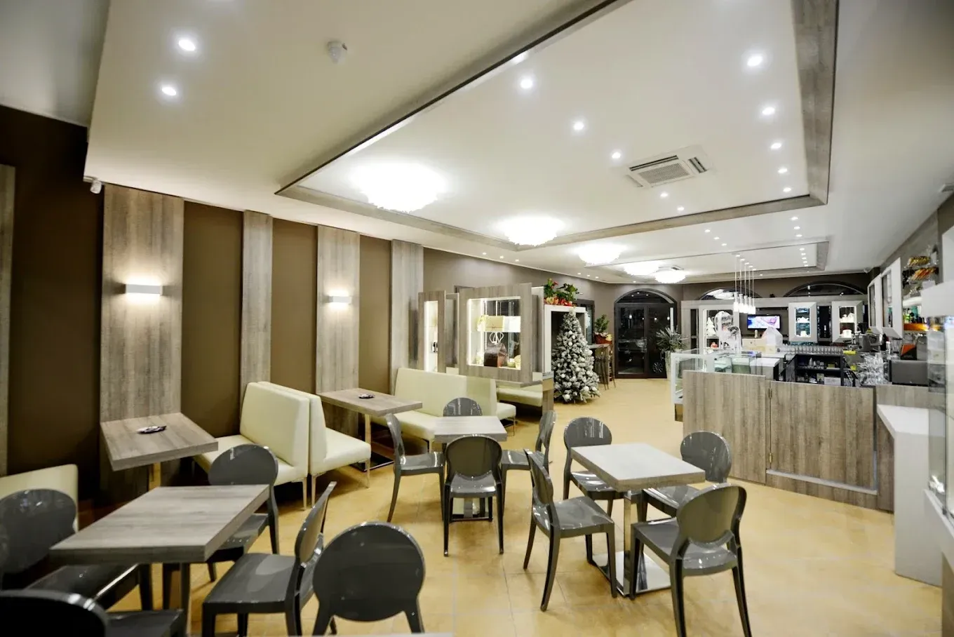 Vista interna di un bar con tavoli, sedie e bancone. Toni neutri, faretti e un albero di Natale sullo sfondo.