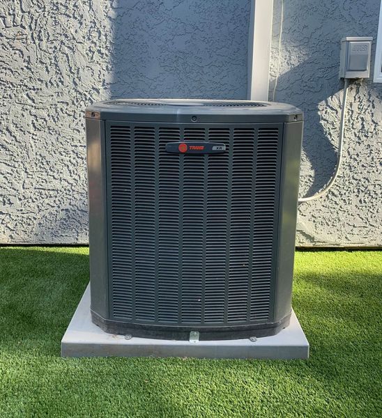 Dark grey Trane air conditioner unit on concrete base, set on green artificial turf. Against stucco wall.