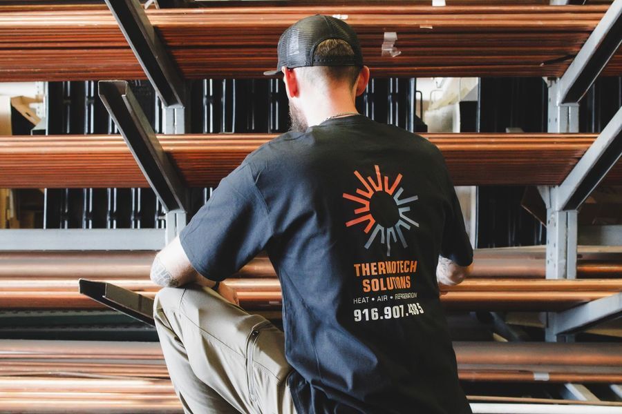 Person in black t-shirt with logo, kneeling in front of metal shelves full of copper tubes.