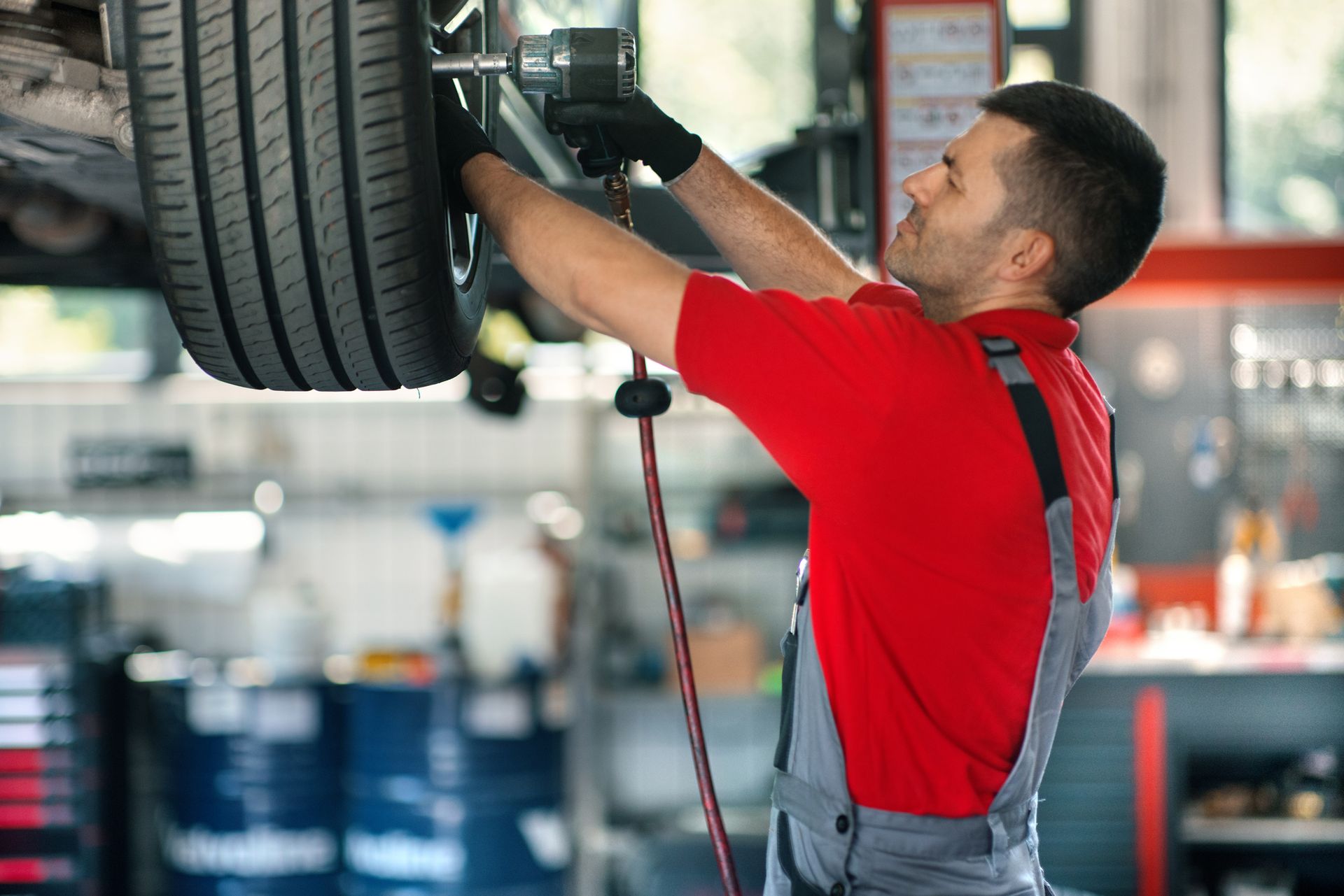 A male car mechanic wearing a red t-shirt and gray overall is replacing a tire at a car shop. A male car mechanic wearing a red t-shirt and gray overall is replacing a tire at a car shop.