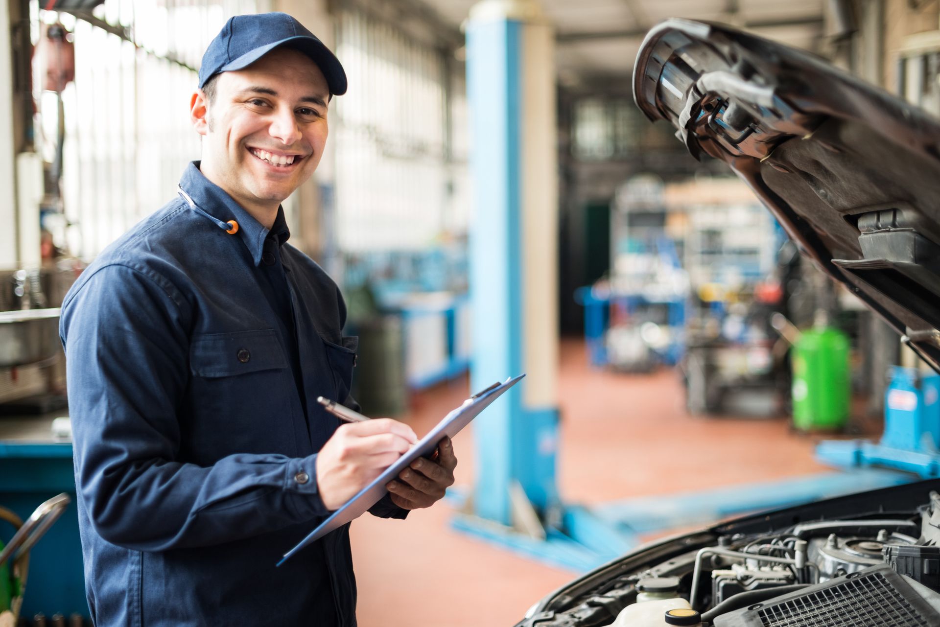 Auto mechanic smiles, holding clipboard, inspecting car engine in a garage.