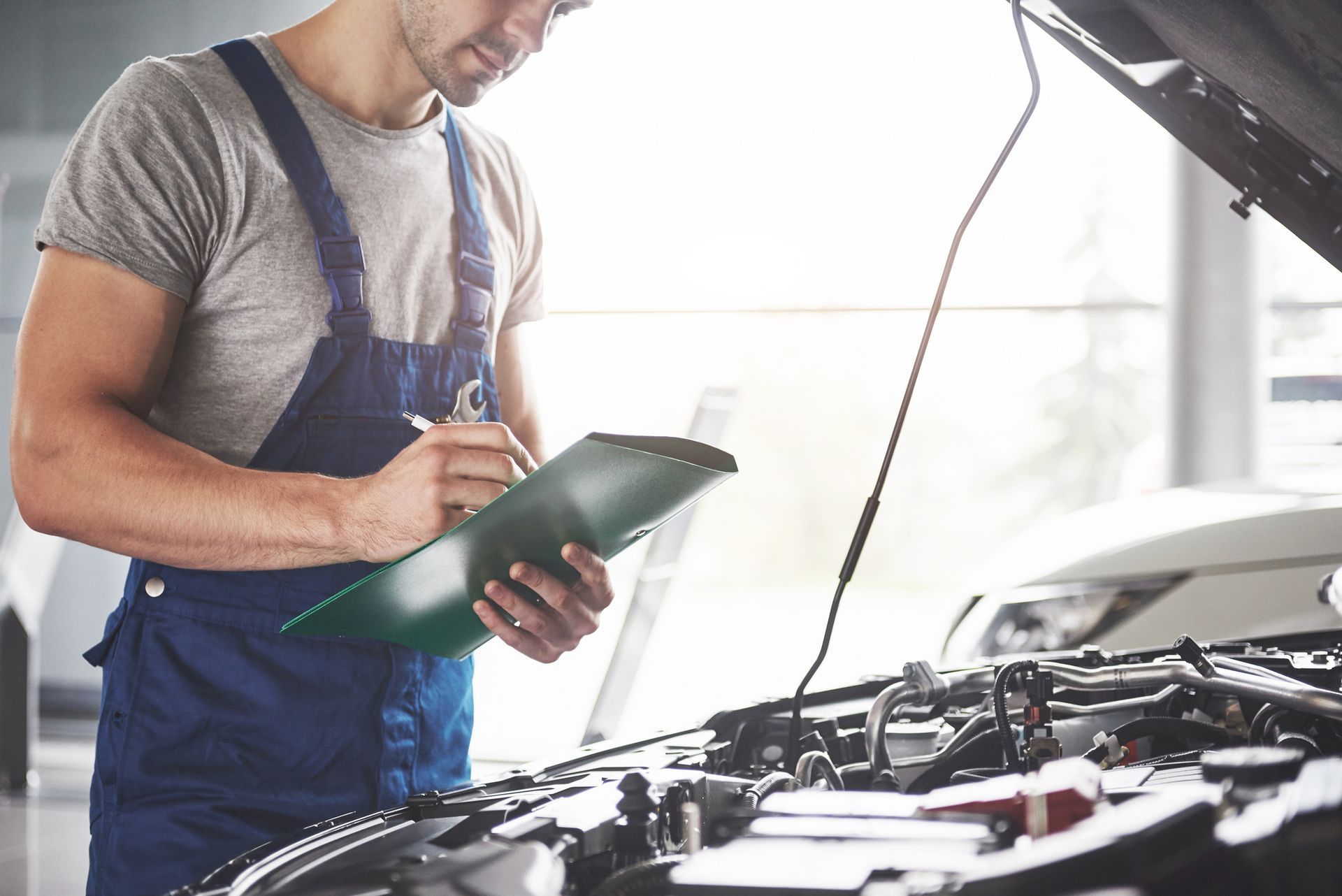 Mechanic in blue overalls examines car engine, writing on clipboard in a garage.