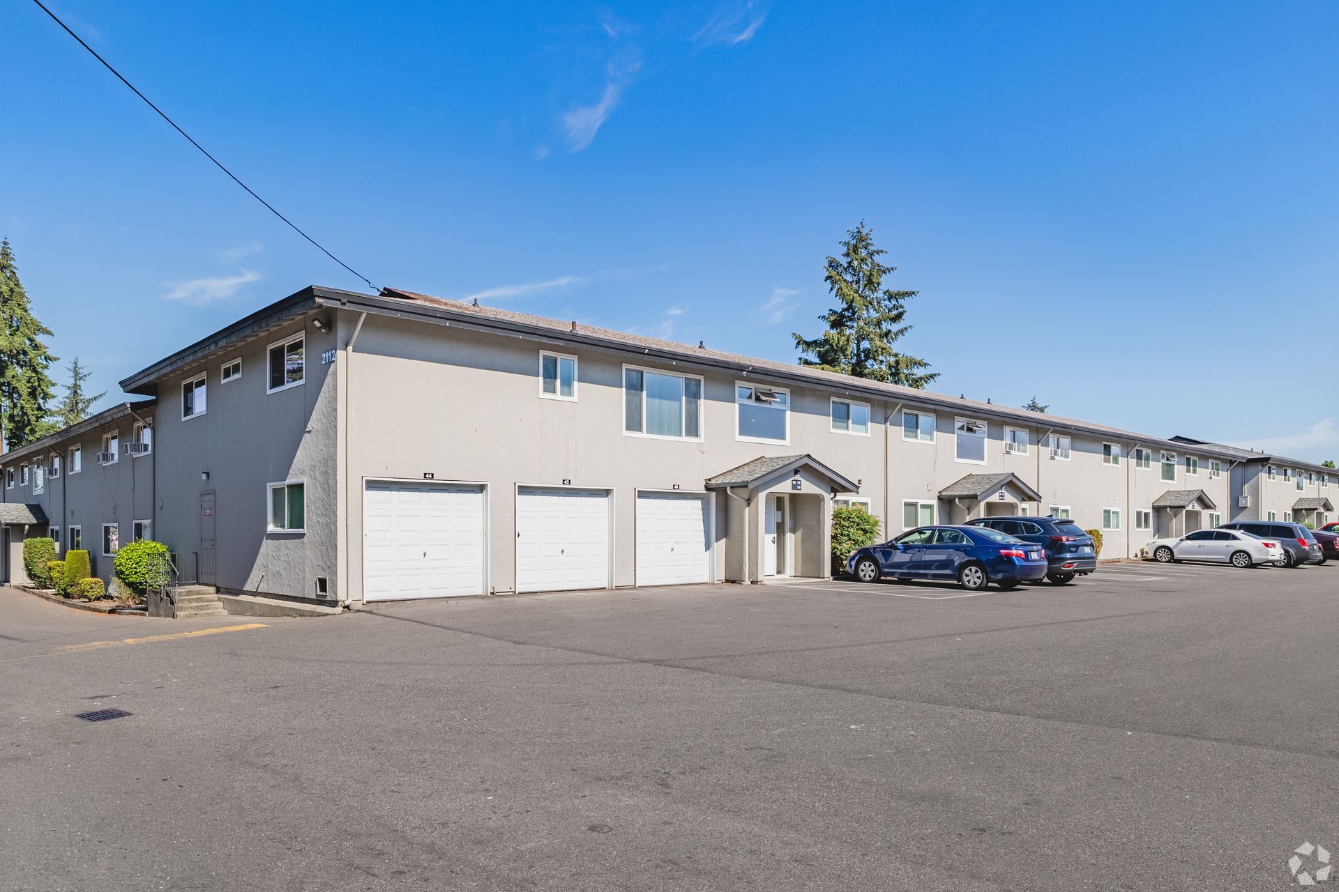Apartment building with garages and parked cars in a paved lot under a clear blue sky