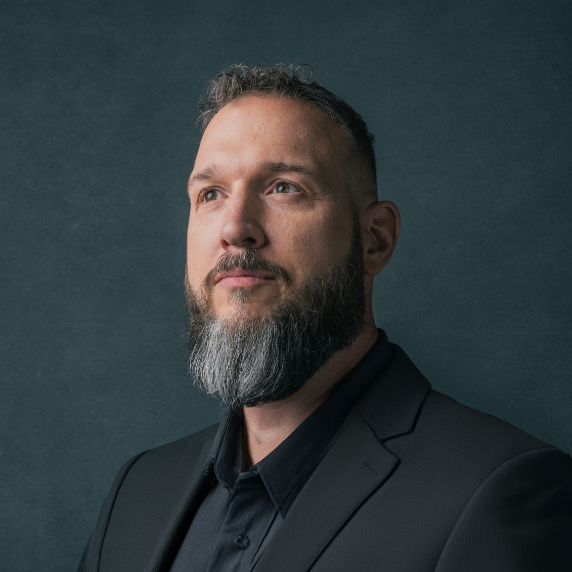 Professional headshot of a bearded man in a dark suit, looking confidently upward against a dark textured background.