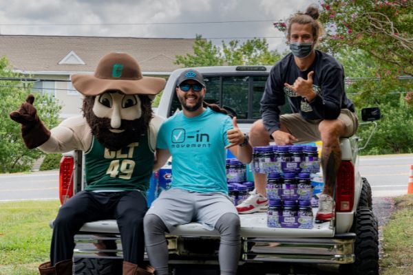 UNC Charlotte mascot, two men, water bottles in truck bed, outdoors. One man in mask gestures, other gives thumbs up.