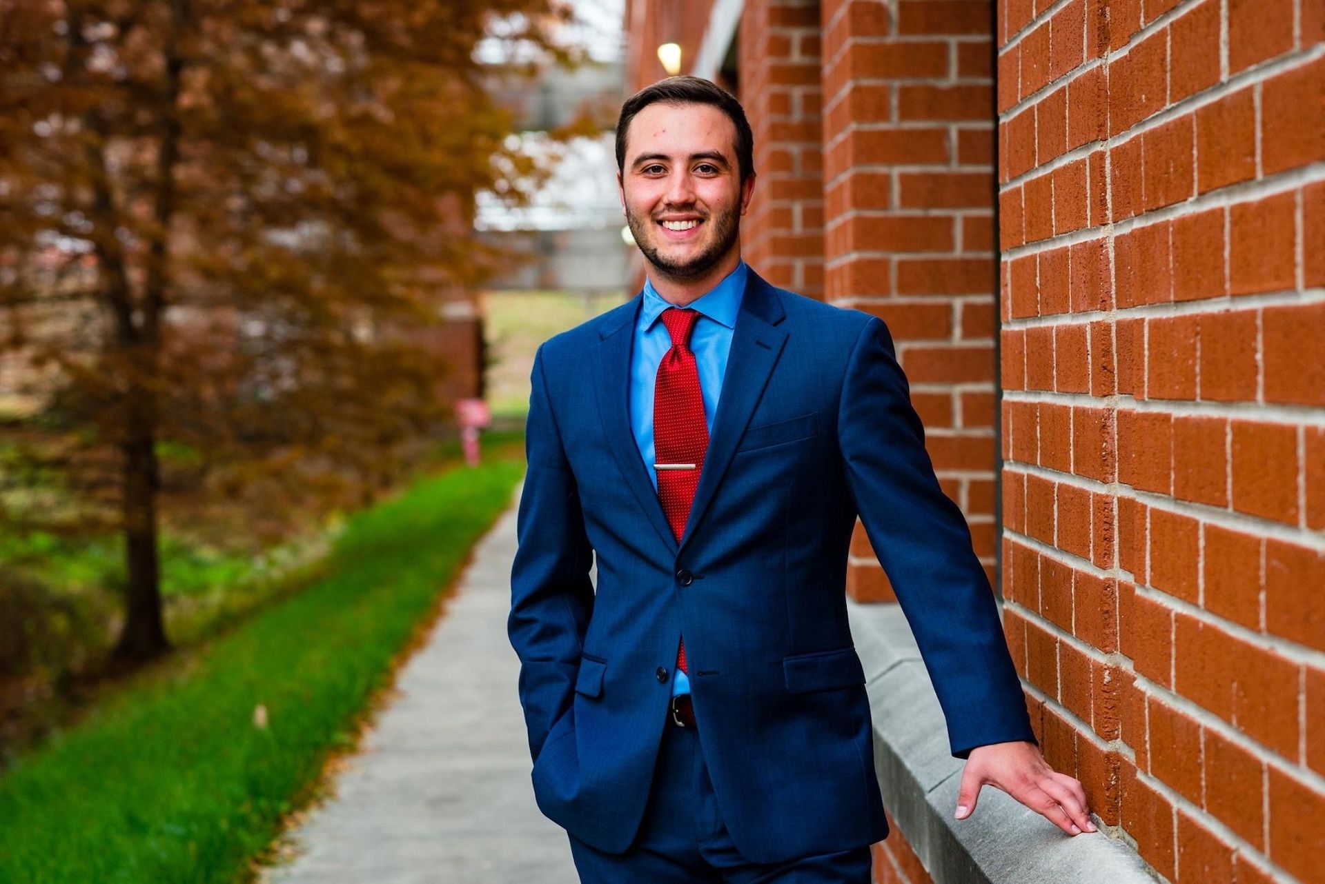 Man in a blue suit and red tie, smiling, leaning on a brick wall. Outdoors with trees.