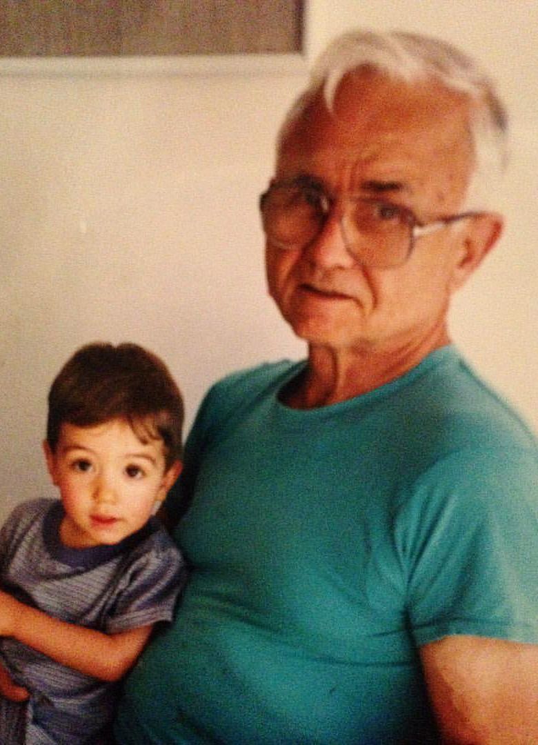 A young child sits with an older man, both looking at the camera. The man wears glasses and a turquoise shirt.