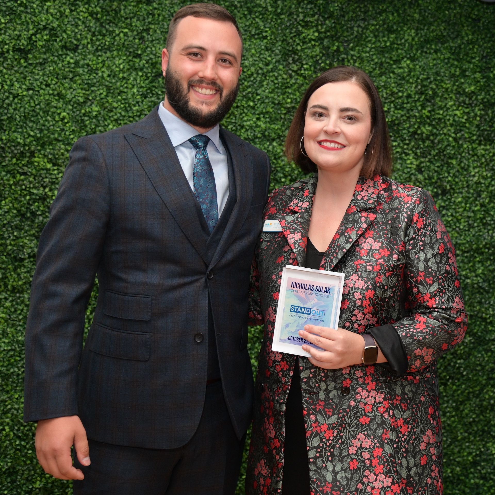 A man and woman pose in front of a green backdrop. The woman holds an award.