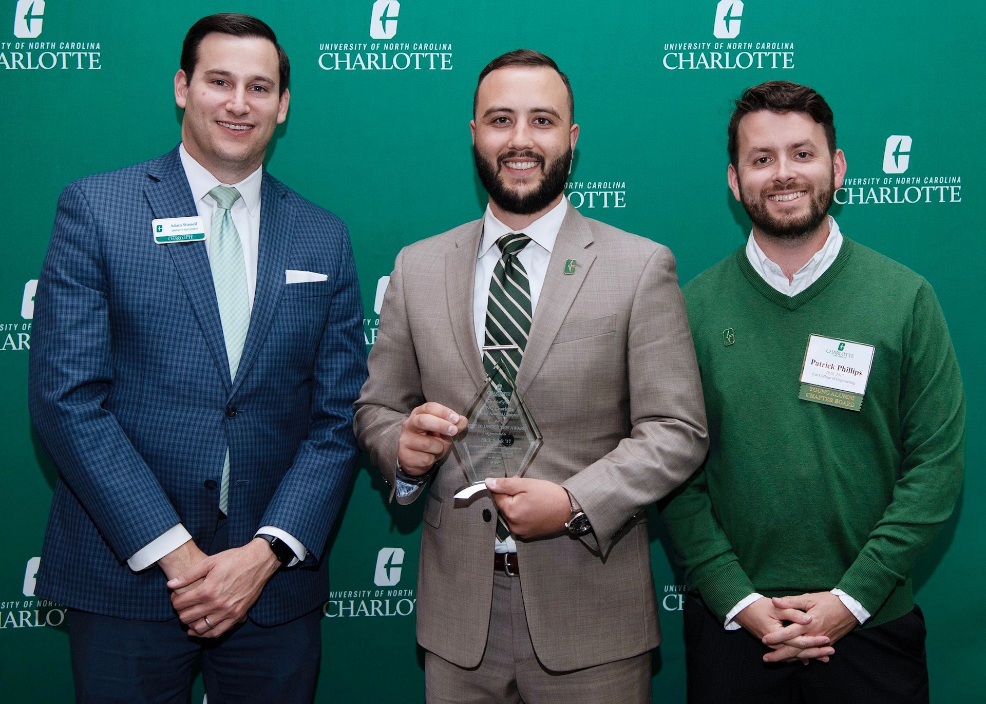Three men posing; one holds an award in front of a green backdrop with a logo.