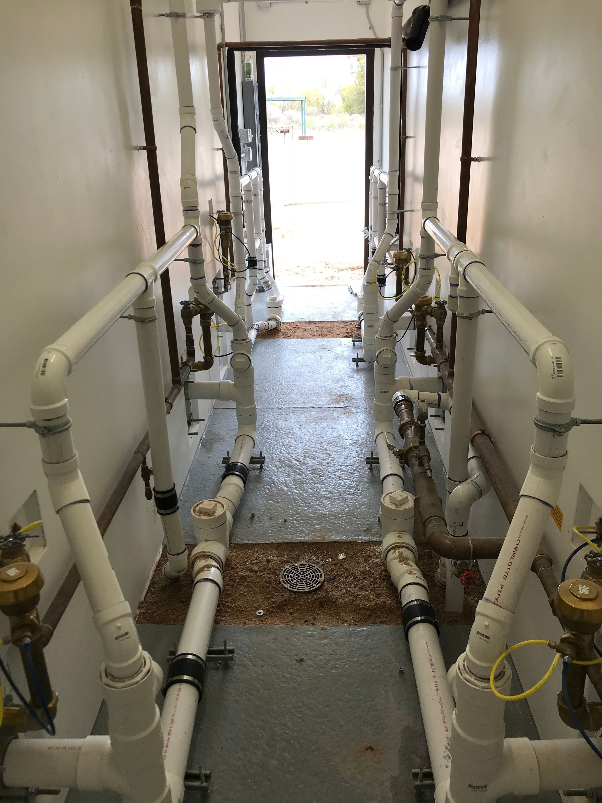 A hallway filled with lots of white pipes leading to a door.