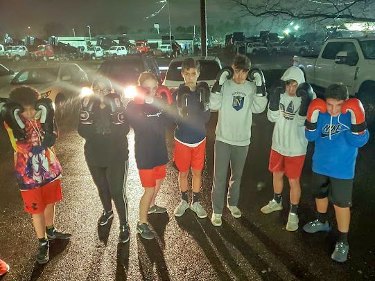 A group of young boys wearing boxing gloves are standing in a parking lot.