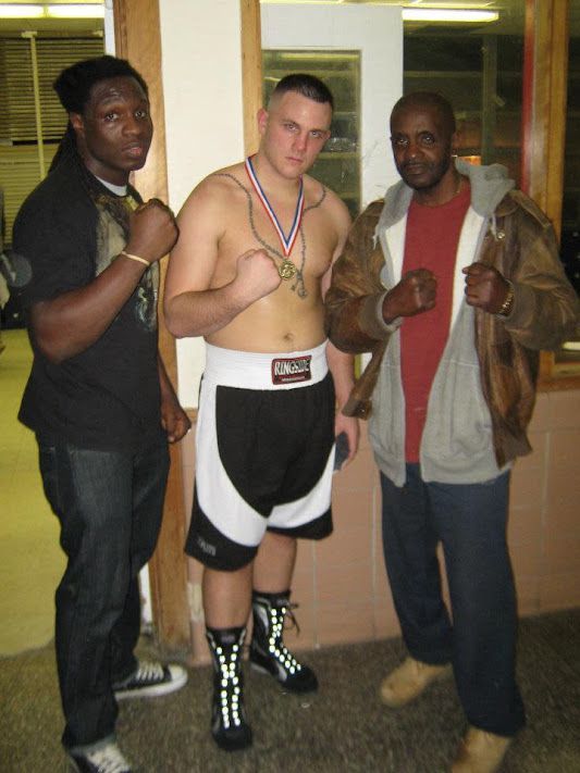 A shirtless boxer with a medal around his neck poses with two other men
