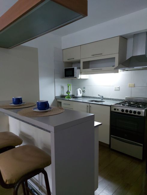 Cocina pequeña con barra de desayuno, dos tazas azules, armarios blancos y una estufa de gas bajo una luz brillante.