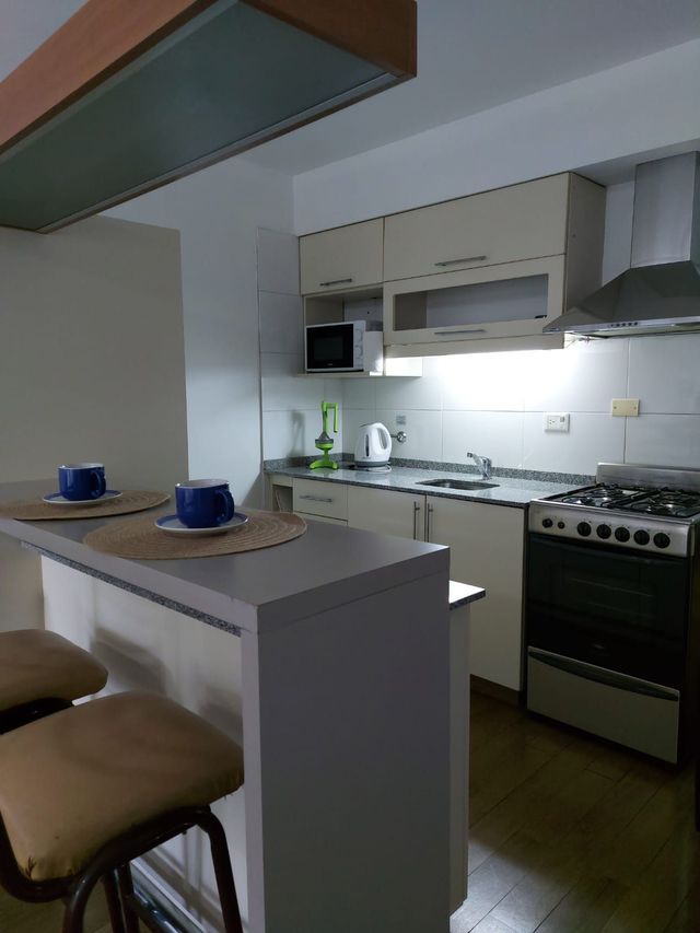 Cocina pequeña con barra de desayuno, dos tazas azules, armarios blancos y una estufa de gas bajo una luz brillante.