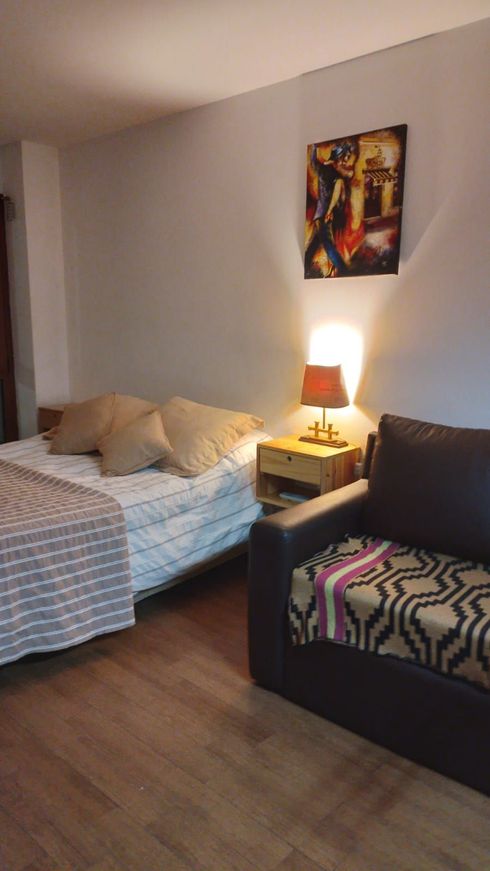 Habitación pequeña con una cama sin hacer, lámpara de noche, cuadros en la pared y un sofá oscuro con cojines estampados.