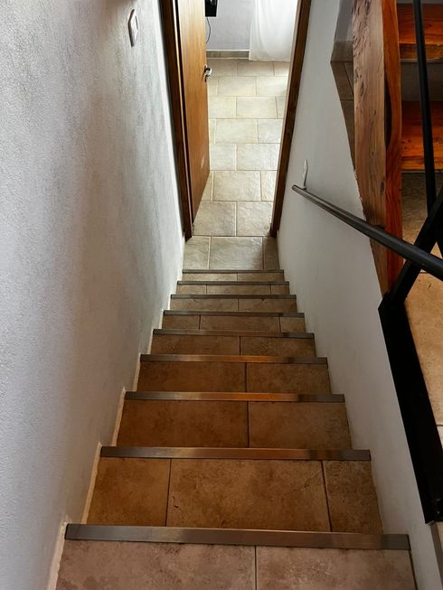 Estrecha escalera interior con escalones de madera y paredes blancas, que conduce a un pasillo alicatado.