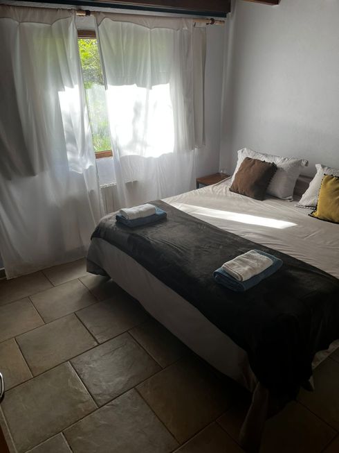 Habitación luminosa con la cama hecha, una manta oscura, almohadas y luz solar que entra a través de cortinas transparentes.
