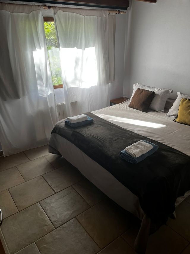 Habitación luminosa con la cama hecha, una manta oscura, almohadas y luz solar que entra a través de cortinas transparentes.