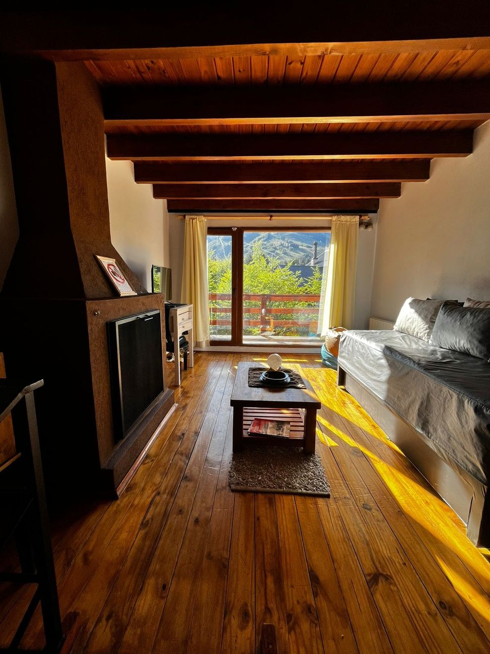 Sala de estar luminosa con vigas de madera, chimenea, sofá y vista a los árboles a través de puertas corredizas de vidrio.