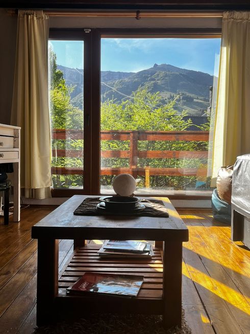 Sala de estar luminosa con vistas a la montaña a través de grandes ventanales, mesa de centro de madera y cortinas.