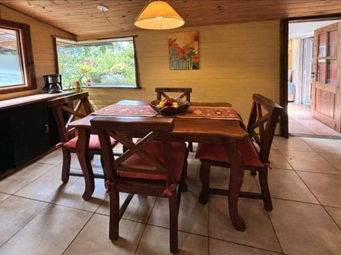 Comedor rústico con mesa de madera, sillas rojas, suelo de baldosas y puerta abierta que da a un patio.