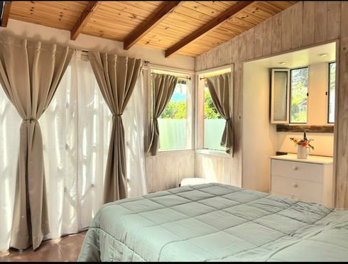 Acogedora habitación con techo de madera, grandes ventanas con cortinas y una cama bien hecha con una colcha azul claro.