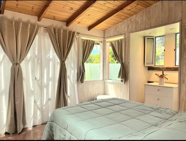 Acogedora habitación con techo de madera, grandes ventanas con cortinas y una cama bien hecha con una colcha azul claro.