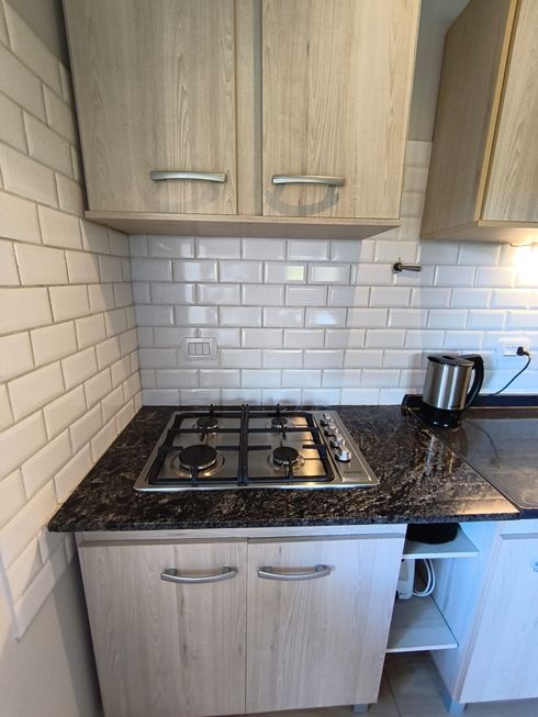 Cocina compacta con gabinetes de madera clara, azulejos blancos tipo metro, encimera negra y placa de gas.