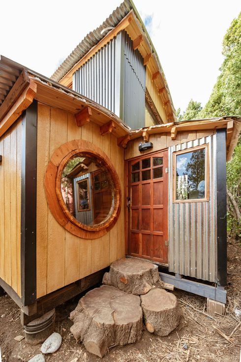 Exterior de una pequeña casa rústica con ventana redonda, revestimiento de madera, techo de metal y escalones de piedra.