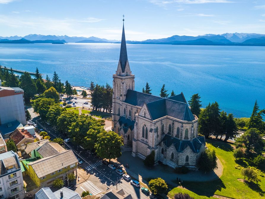 Iglesia de piedra con una alta aguja junto a un lago azul, enmarcada por árboles y montañas bajo un cielo despejado.
