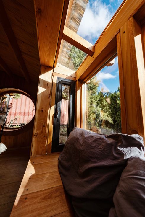 Pasillo de madera iluminado por el sol con tragaluz, puerta abierta y sillón tipo puf dentro de una casa pequeña moderna.