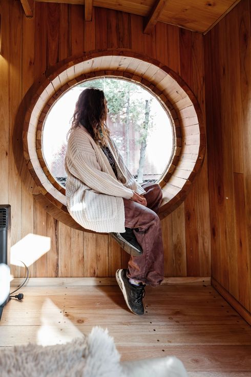 Persona sentada junto a una ventana redonda en una habitación de madera, con un abrigo claro estampado y botas marrones.