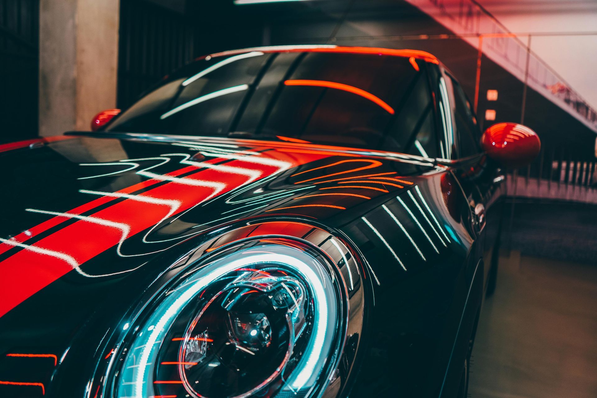 Black car with red stripe, illuminated headlights, parked indoors, reflective surfaces.