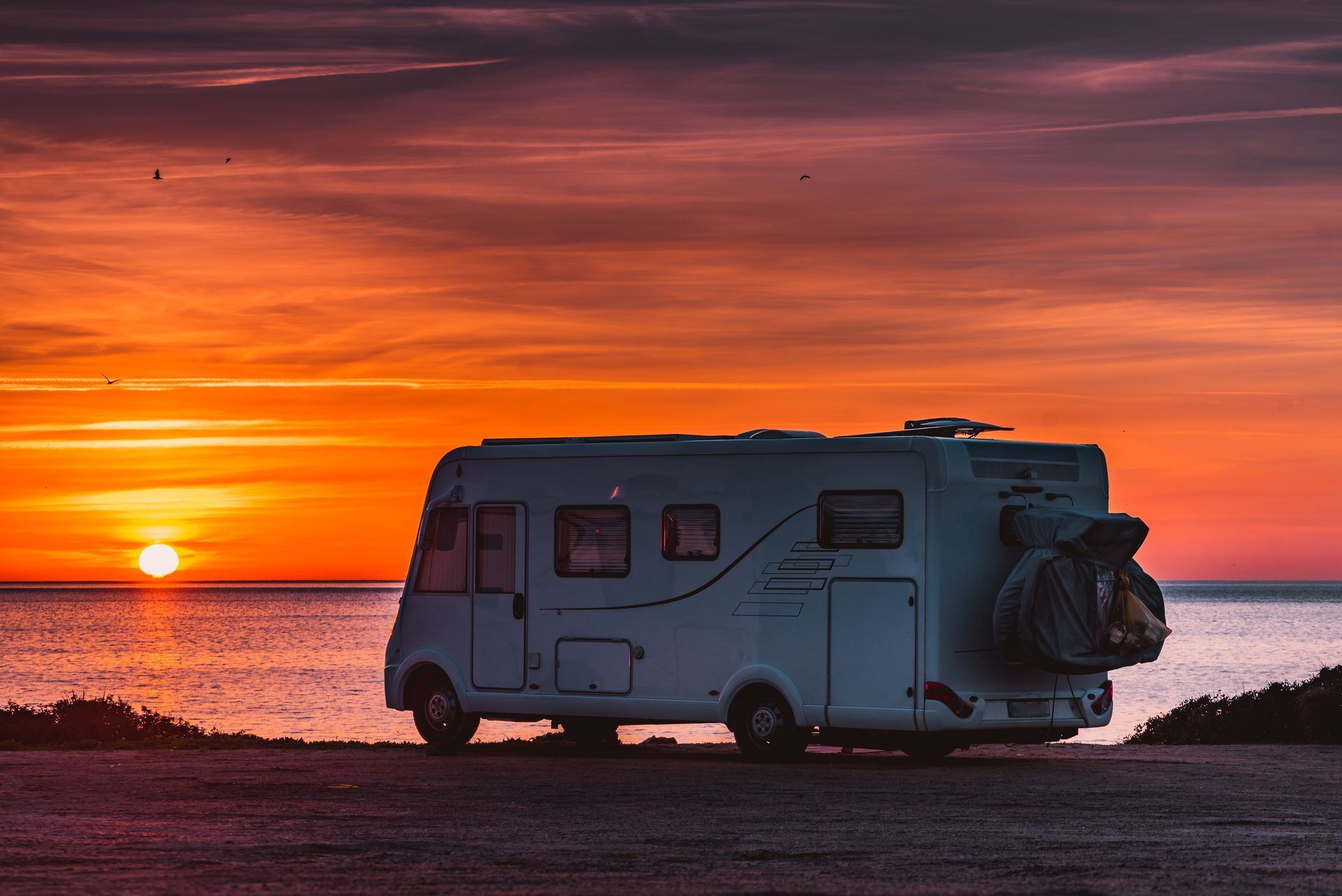 RV parked on a beach watching a vibrant sunset over the ocean; orange, red, and yellow hues.