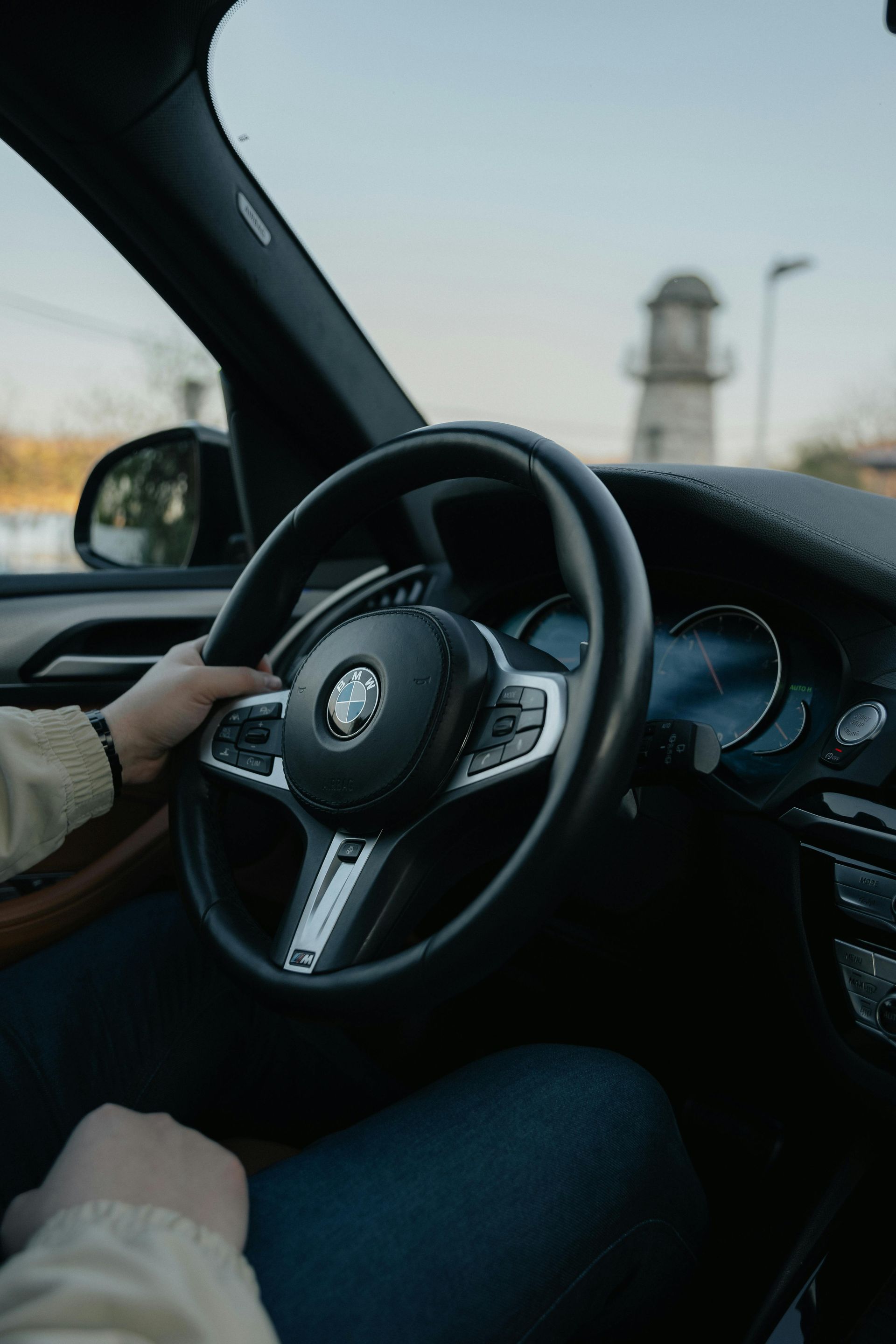 Person's hands on BMW steering wheel, driving. Black interior. Tower visible outside.