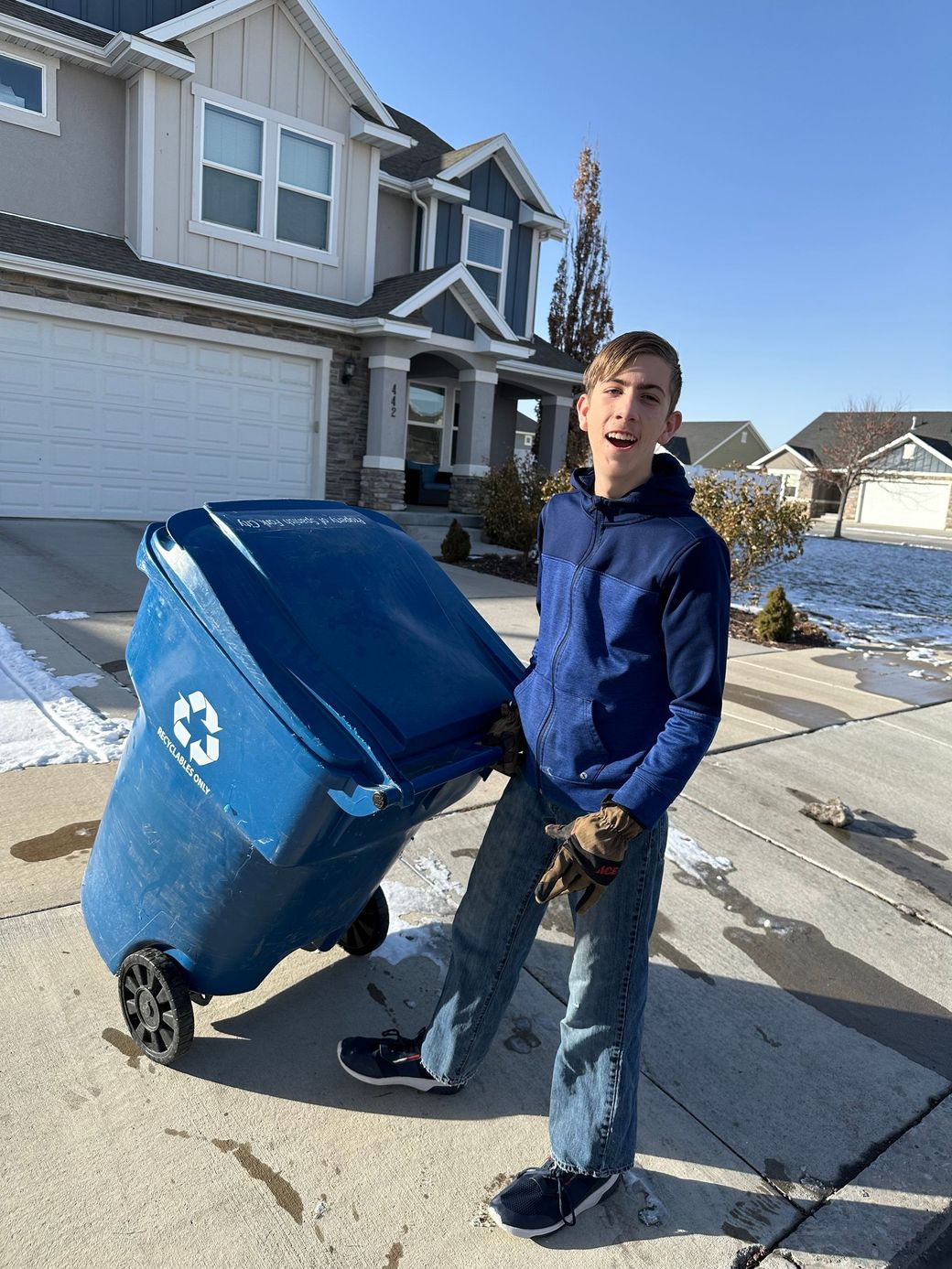 A young man is pushing a blue trash can down a sidewalk in front of a house.