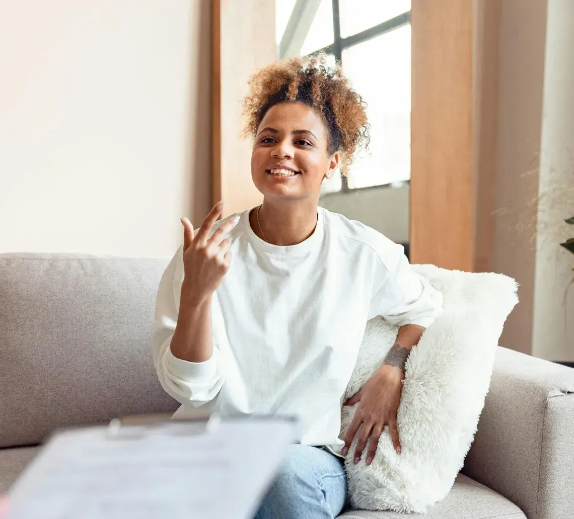 A person speaks while gesturing during a meeting, seated on a gray sofa across from someone holding a clipboard.
