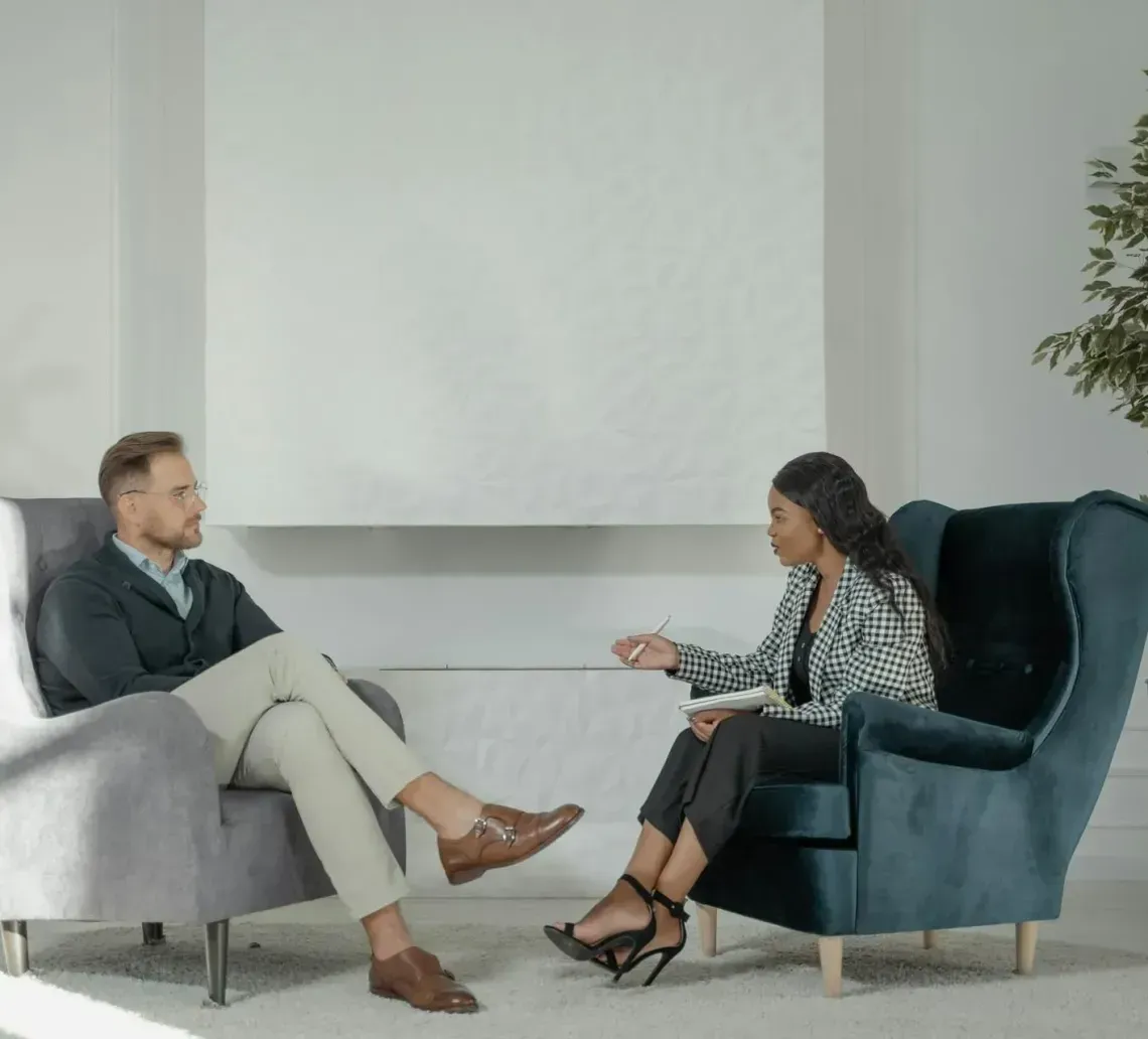 A person in a dark cardigan and light trousers sits in a gray chair, speaking with someone in a patterned blazer.